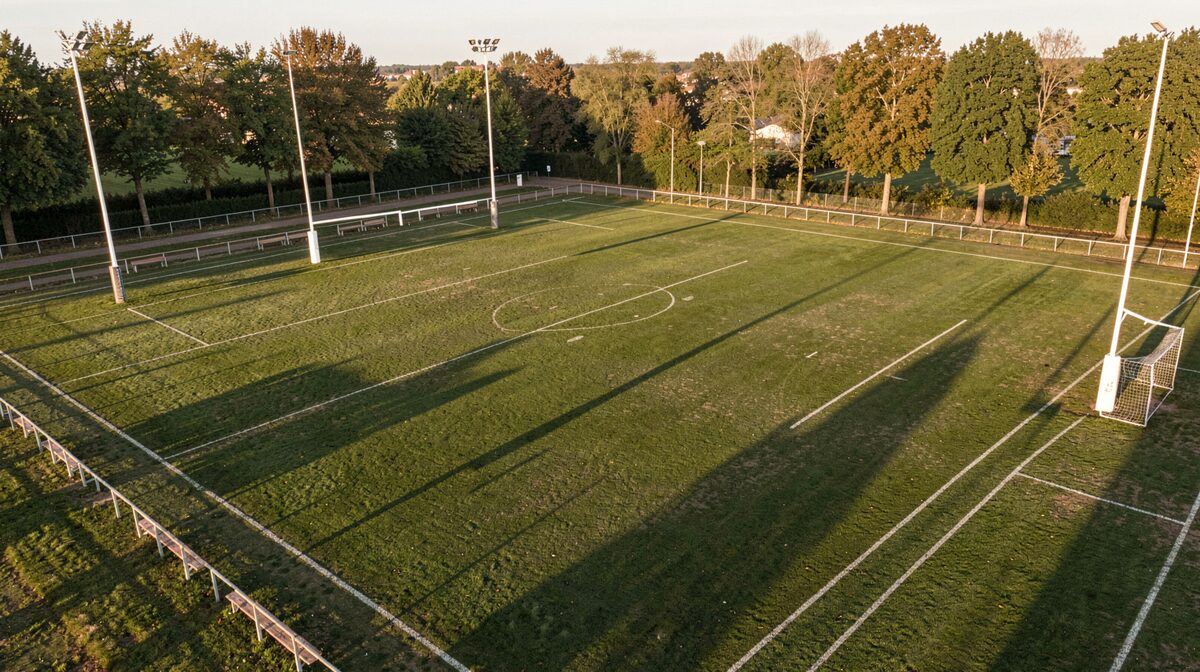 Vue en plongée d'un terrain de rugby municipal en Gironde en fin d'après-midi, poteaux blancs et lignes tracées à la chaux, bancs de touche vides et ombre longue des arbres en bord de terrain, lumière rasante dorée.