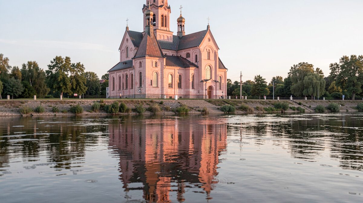Une cathédrale de brique rose vue depuis une rive, lumière rasante du matin, cadrage large avec le reflet dans l'eau au premier plan.
